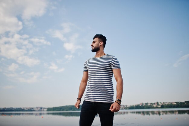 Handsome tall arabian beard man model at stripped shirt posed outdoor against lake and sky Fashionable arab guy