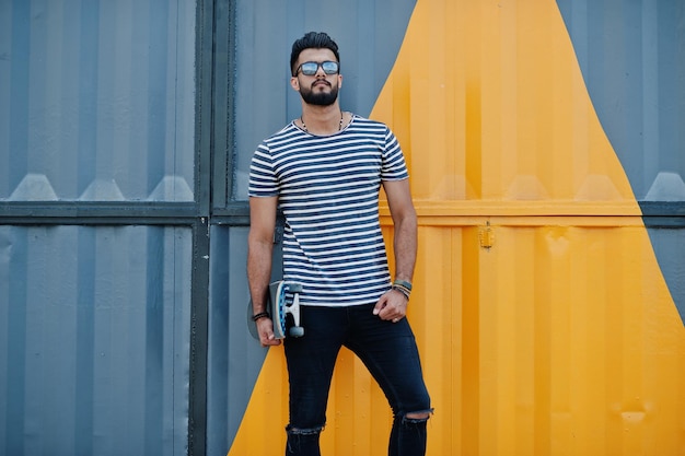 Handsome tall arabian beard man model at stripped shirt posed outdoor Fashionable arab guy at sunglasses with skateboard against yellow painted wall
