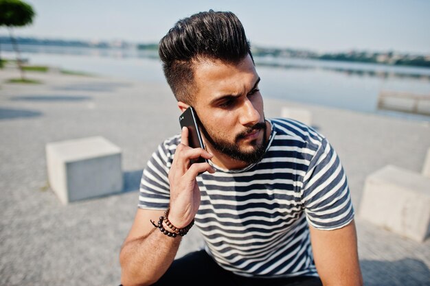 ストリップシャツでハンサムな背の高いアラビアのひげの男のモデルは、携帯電話で屋外のファッショナブルなアラブの男を提起しました