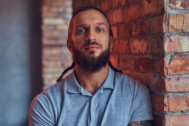 A handsome tattoed male with a stylish haircut and beard, in a gray t-shirt, standing leaning against a brick wall in a room with a loft interior.