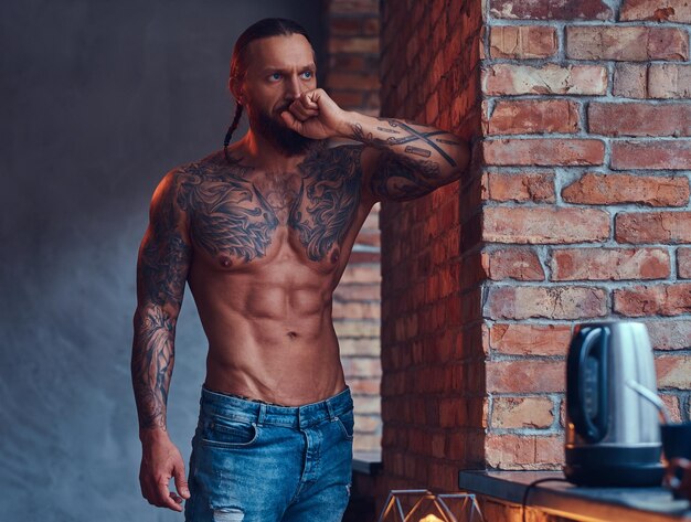 Красивый татуированный мужчина без рубашки со стильной стрижкой и бородой, стоящий, прислонившись к кирпичной стене на кухне.