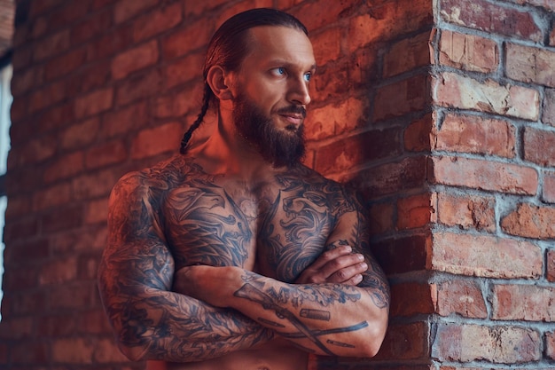 Handsome tattoed shirtless male with a stylish haircut and beard, standing with crossed arms, leaning against a brick wall in a room with a loft interior.