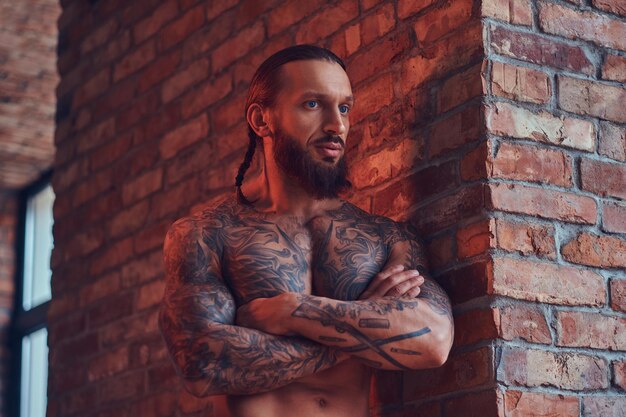 Handsome tattoed shirtless male with a stylish haircut and beard, standing with crossed arms, leaning against a brick wall in a room with a loft interior.