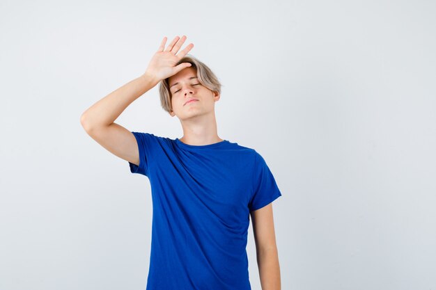 Handsome teen boy in blue t-shirt keeping hand on head, shutting eyes and looking tired , front view.