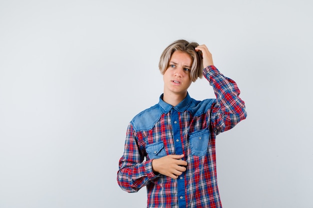 Handsome teen boy in checked shirt scratching head, looking away and looking concerned , front view.