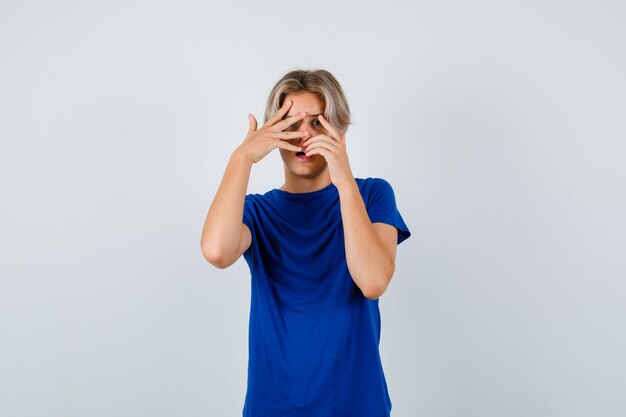 Handsome teen boy peeking through fingers in blue t-shirt and looking frightened. front view.