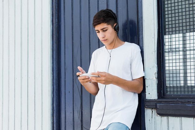 Красивый подросток, опираясь на металлическую стену с помощью смартфона и прослушивания музыки