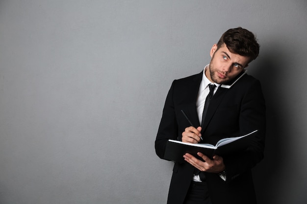 Handsome thinking businessman taking notes while talking on mobile phone, lookin aside