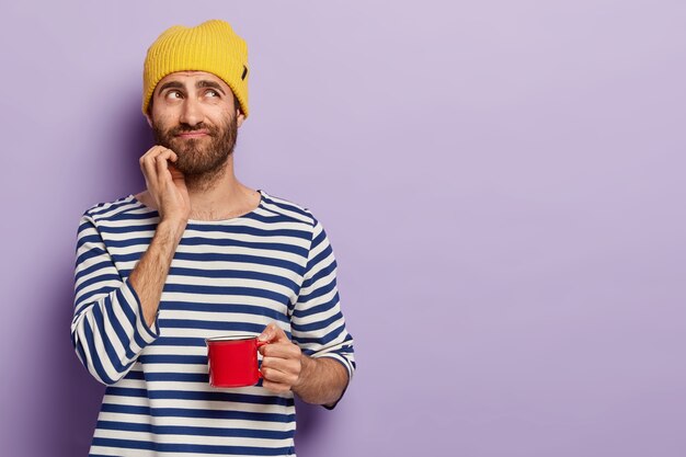 Handsome thoughtful young man with bristle, holds mug of coffee, has pensive look, break after work, wears striped jumper, yellow headgear