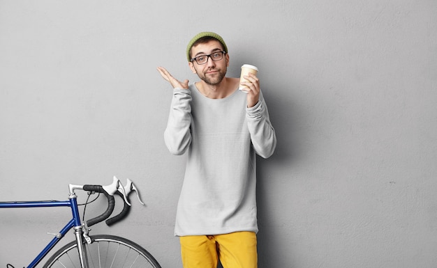 Bel turista a riposo per un po 'dopo aver guidato in bicicletta in luoghi sconosciuti, bevendo caffè dalla tazza di carta, alzando le spalle con incertezza, non conoscendo la sua prossima destinazione. persone e stile di vita