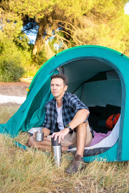 Handsome tourist sitting in tent and looking on scenery. Thoughtful Caucasian traveler camping on nature and drinking tea from vacuum flask. Backpacking tourism, adventure and summer vacation concept