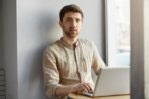 검은 머리가 창 근처의 coworking office에서 일하고, 사려 깊은 표정으로 옆을보고,해야 할 일을 기억하려고하는 잘 생긴 형태가없는 사람.