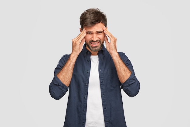 Handsome unshaven male keeps hands on temples, frowns face in displeasure, has terrible headache, dressed in white blue shirt, poses against white wall. Puzzled discontent man poses indoor