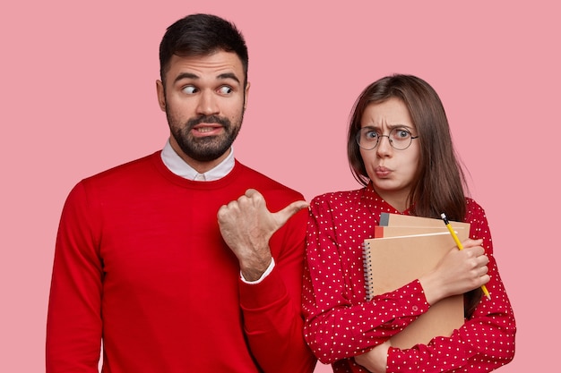 Handsome unshaven man wears red jumper, points at displeased beautiful woman who makes grimace, carries orgnaizer and spiral notepad