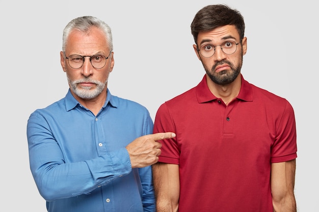 Handsome unshaven serious serious male in spectacles and formal shirt indicates at his clueless colleague, being experienced, frowns face with puzzlement, wears red t-shirt, stand together indoor