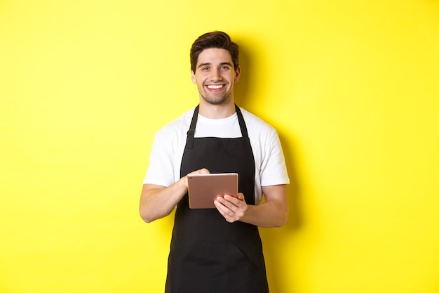 注文を受けて、デジタルタブレットを持って、笑顔で、黒いエプロンのユニフォームを着て、黄色の背景の上に立っているハンサムなウェイター。