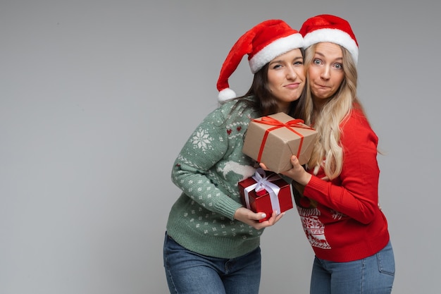 Handsome women friends in red and white christmas hats holds a presents for each other and smiles