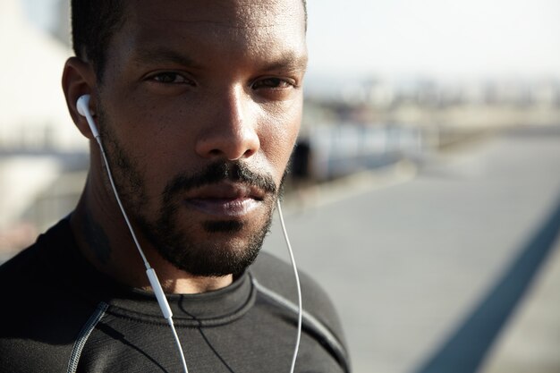 Handsome young African American runner or jogger wearing sportswear exercising in open air in the morning. Attractive black male listening to motivative music for training using his earphones