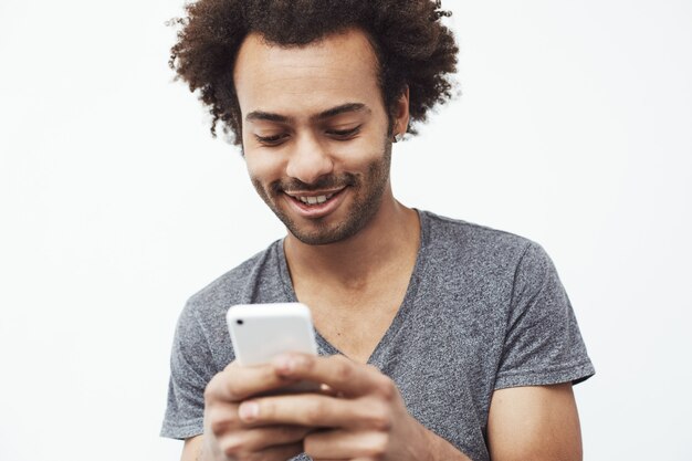 Handsome young african male surfing the web on his smartphone, browsing social media, posting on sites over white wall.