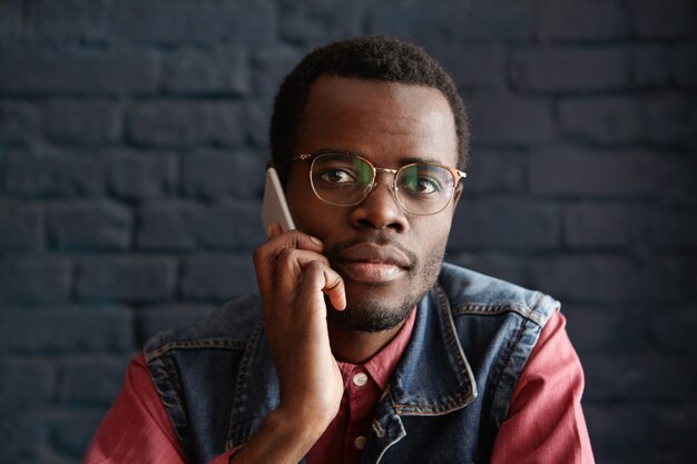 Handsome young African male wearing stylish glasses talking on mobile phone with his girlfriend, waiting for her