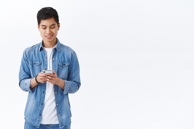 Handsome young asian male student texting friend, holding smartphone, stay connected using mobile phone, internet, scroll social network online, white wall