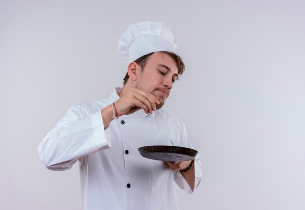 Красивый молодой бородатый шеф-повар в белой униформе и шляпе держит сковороду на белой стене