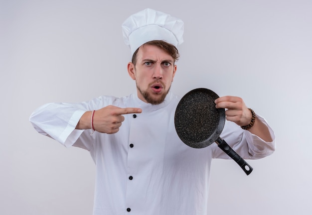 Un bel giovane chef barbuto uomo che indossa l'uniforme bianca del fornello e cappello che punta alla padella mentre guarda con espressione aggressiva su un muro bianco