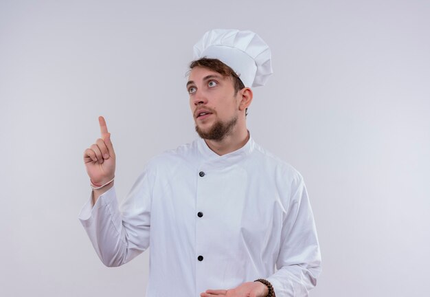 A handsome young bearded chef man wearing white cooker uniform and hat pointing up with index finger on a white wall