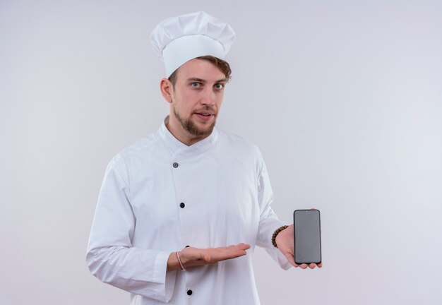Красивый молодой бородатый шеф-повар в белой униформе и шляпе показывает пустое место мобильного телефона на белой стене