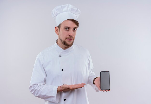 A handsome young bearded chef man wearing white cooker uniform and hat showing blank space of mobile phone on a white wall