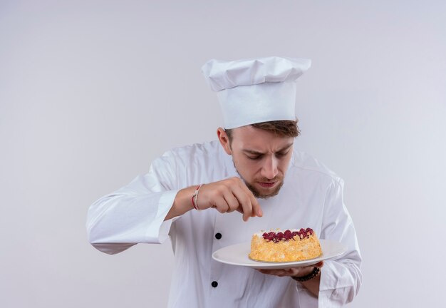 A handsome young bearded chef man wearing white cooker uniform and hat touching a plate with cake on a white wall