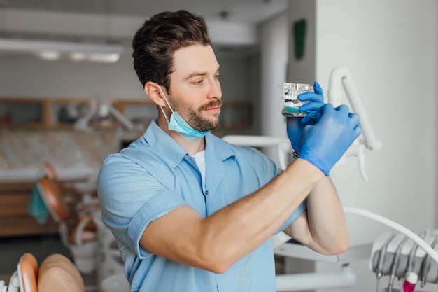 Handsome young bearded dentist in white coat is holding plastic layot and toothbrush, while standing in his office.