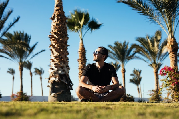 Handsome young bearded man in sunglasses sitting on the grass under palms on summer vacation luxary resort