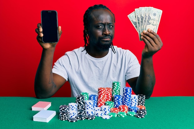 Красивый молодой темнокожий мужчина, играющий в покер со смартфоном и долларами, скептически и нервно хмурится из-за проблемного негативного человека