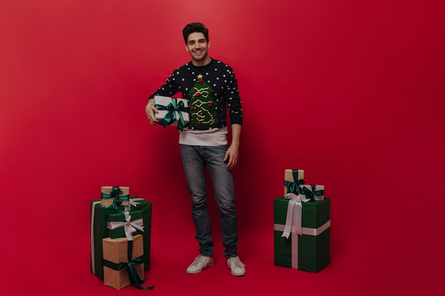 Handsome young brunette in christmas sweater blue jeans and light sneakers smiling looking into camera and posing with many gift boxes against plain red background