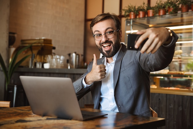 Handsome young businessman dressed in suit working on a laptop computer, sitting at the cafe indoors, taking a selfie