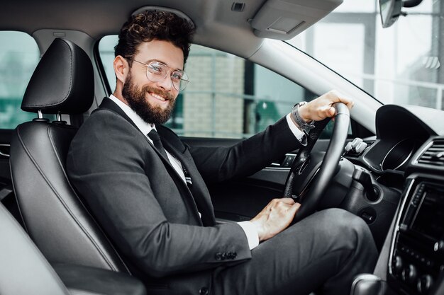 Handsome young businessman in full suit smiling while driving a new car