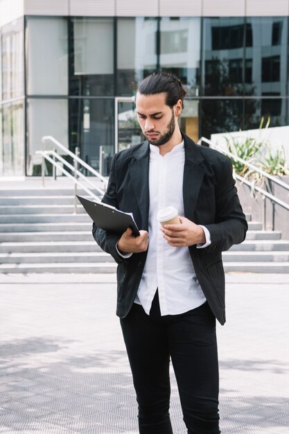 Handsome young businessman holding disposable paper cup looking at clipboard