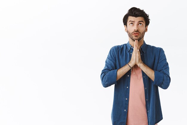 Handsome young caucasian bearded guy praying god look up in sky and making wish clasp hands together over chest supplicating beggingr Lord for good new year standing white background