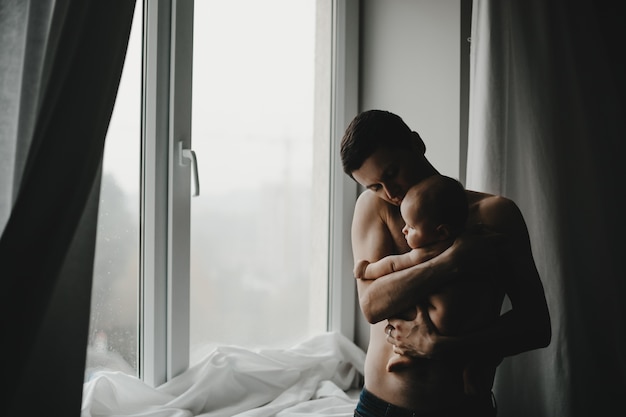 Free photo handsome young dad holds newborn baby close to his heart standing before a window