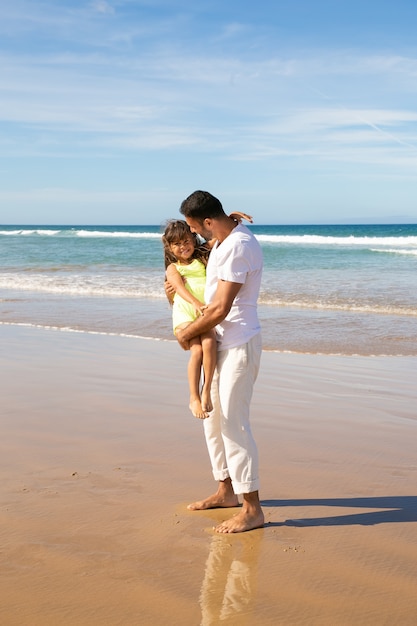 海のビーチで小さな娘と余暇を過ごし、子供を腕に抱いてハンサムな若いお父さん
