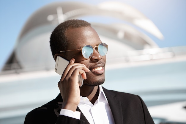 Handsome young dark-skinned businessman in trendy mirrored lens shades and formal suit holding mobile phone, having conversation with his partner, sharing great news concerning business issues