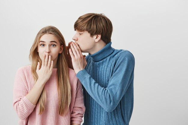 Handsome young fair-haired male student in blue sweater, whispering something into ear of stylish blonde girl