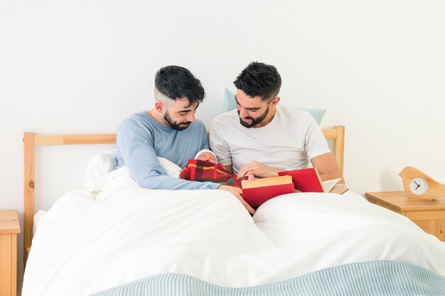 Giovani coppie gay belle con i libri a disposizione che guardano il loro bambino avvolto in coperta