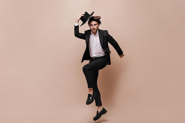 Free photo handsome young gentleman with dark hair glasses light shirt and black striped suit jumping posing on one leg taking off his hat and looking straight against beige background