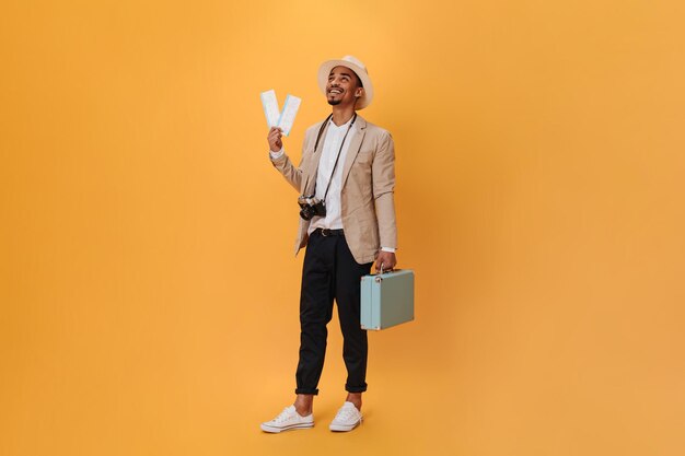 Handsome young guy posing in studio on isolated Charming happy man in black pants and beige jacket posing with suitcase and tickets