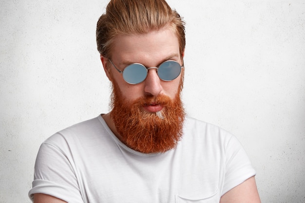 Handsome young hipster guy, has stylish hairdo, red beard and mustache, wears trendy sunglasses, dressed in white t shirt, isolated over white concrete