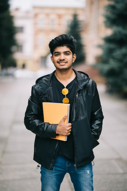 Handsome young indian student man holding notebooks while standing on the street