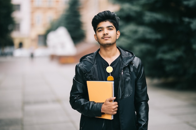 Free photo handsome young indian student man holding notebooks while standing on the street