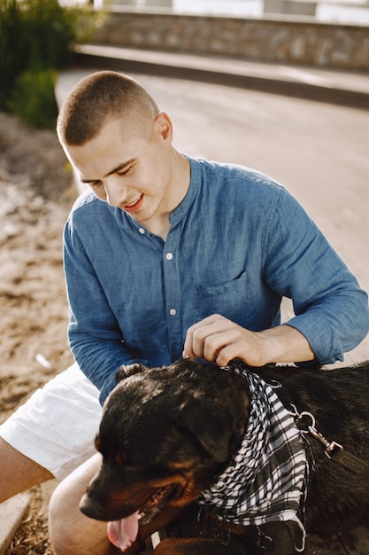 Foto gratuita bel giovane maschio in abito casual che gioca con un cane carino mentre è seduto vicino al lago. ragazzo che indossa una maglietta blu e pantaloncini di jeans bianchi